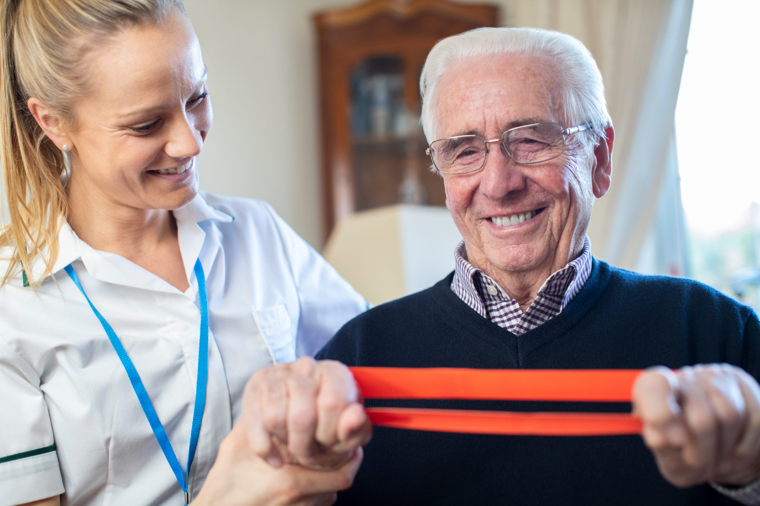 Female Physiotherapist Helping Senior Man To Use Resistance Band At Home