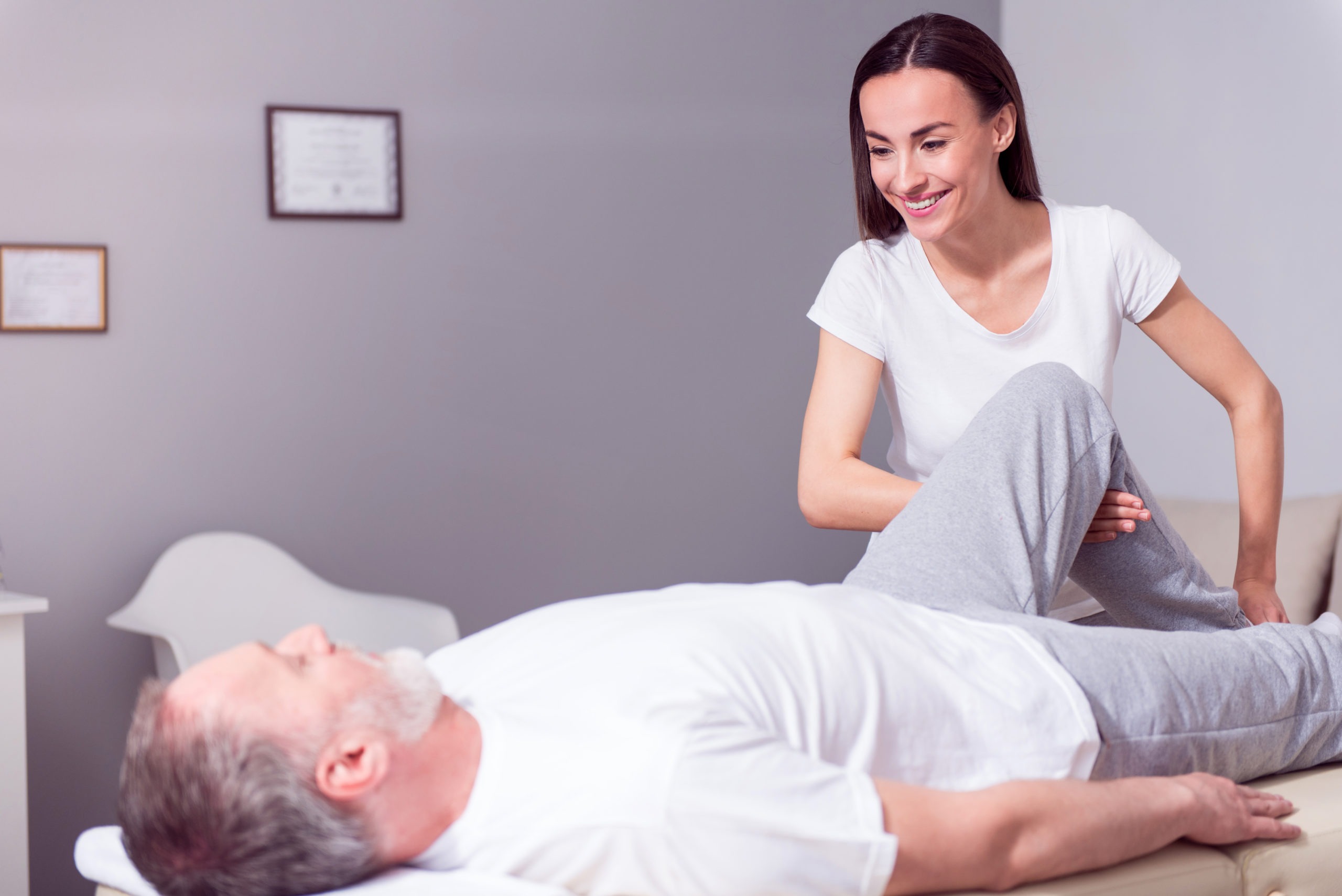 Professional help. Glad male patient lying down with female positive physiotherapist performing some stretch exercises on mans leg